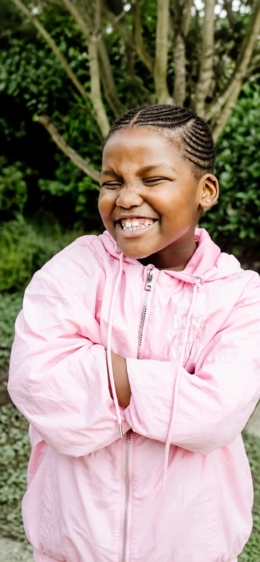 A JENS learner in a pink jacket laughing with her arms folded outside in the school garden