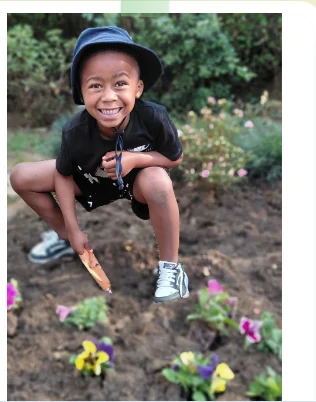 A beaming JENS learner in a sunhat crouching with a trowel beside a bed of freshly planted yellow and purple pansies