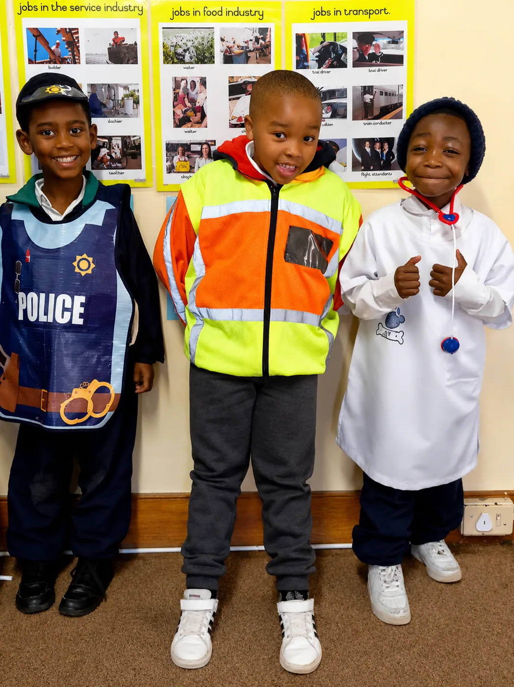 Three JENS Grade R learners dressed as a police officer, a road worker and a doctor in front of the classroom careers posters