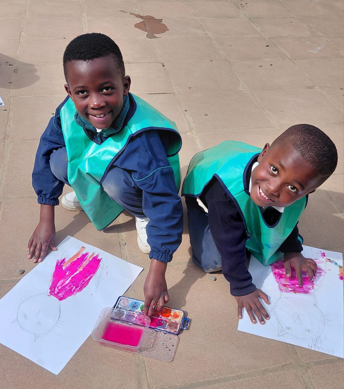 Two JENS Grade 1 learners in green aprons kneeling outdoors, dipping their fingers into pink paint beside their cat drawings
