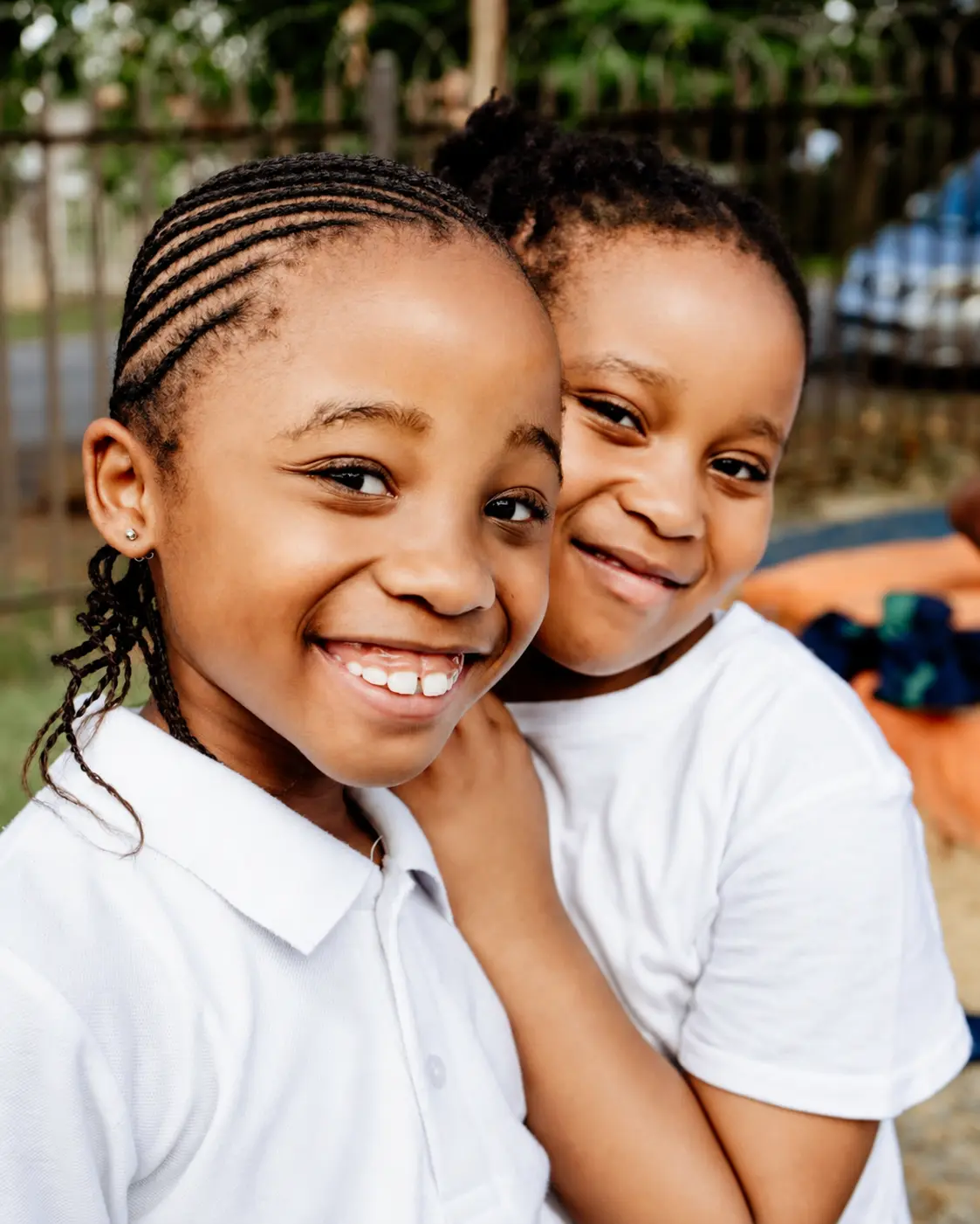 Two JENS learners in white school shirts hugging cheek to cheek and smiling
