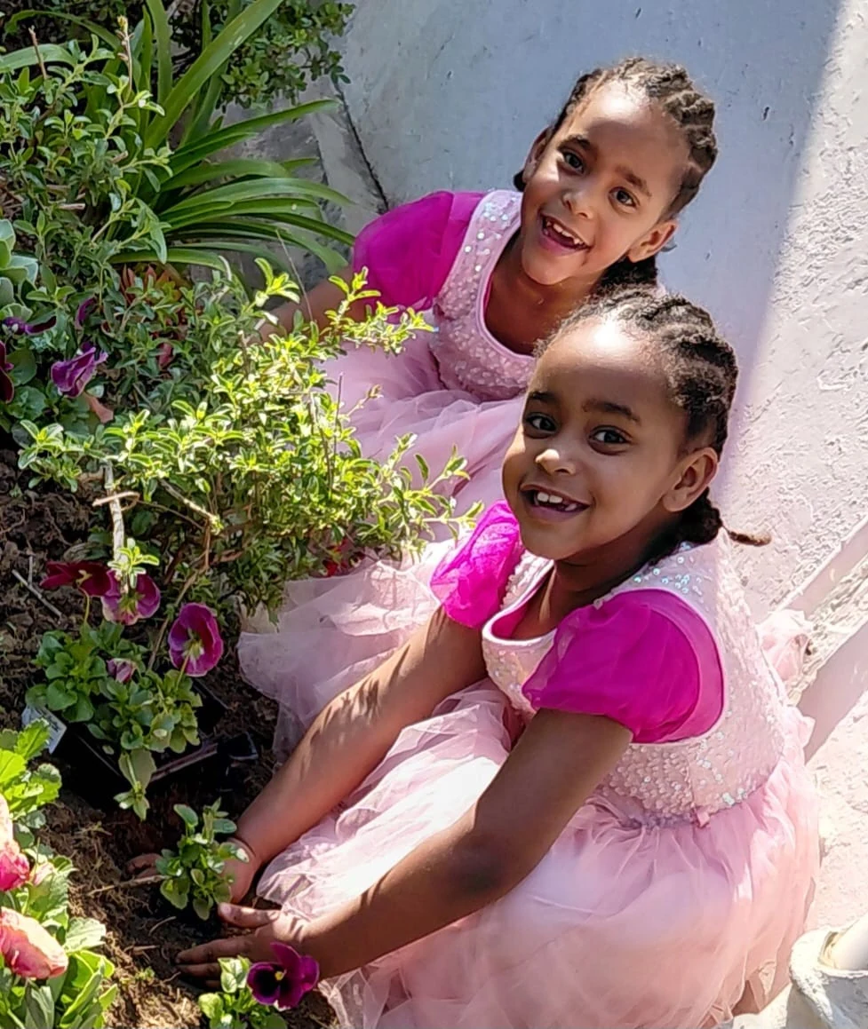 Two JENS learners in pink dresses kneeling together to plant seedlings in a flower bed