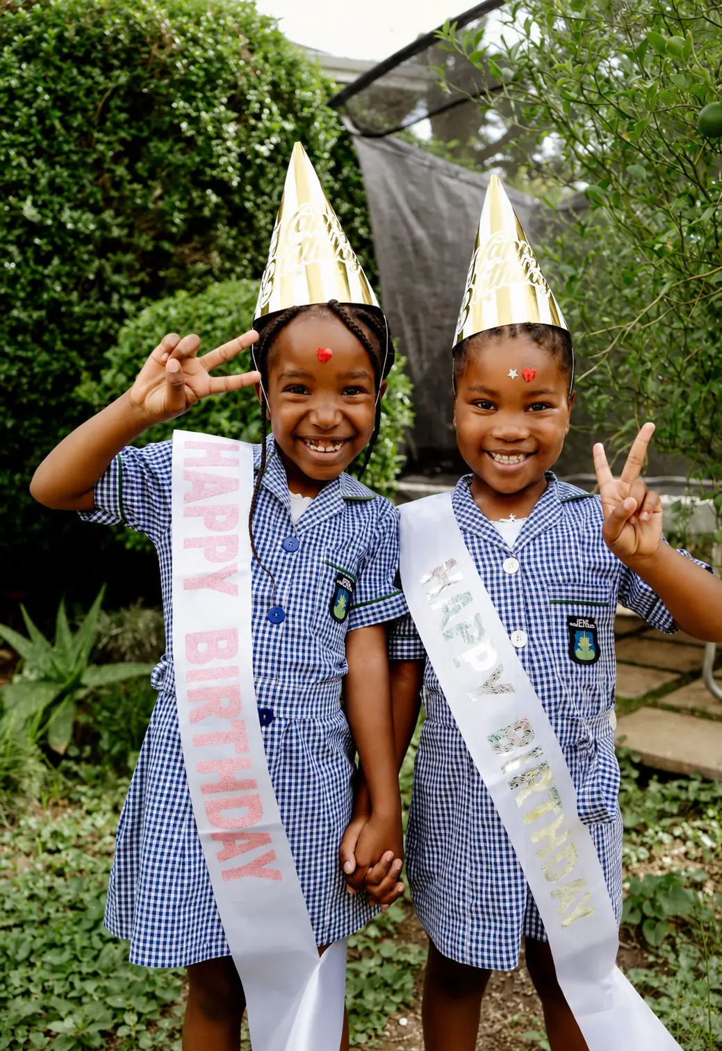 Two JENS learners in checked school dresses wearing birthday hats and sashes, holding hands and smiling