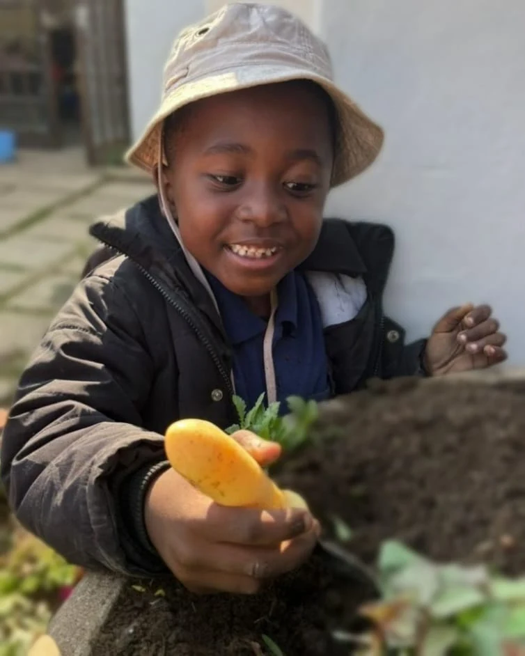 A smiling JENS learner in a sunhat holding up a freshly dug potato beside a garden bed on Spring Gardening Day