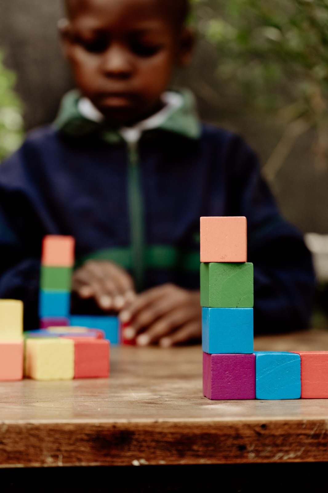 A JENS learner in a navy school jacket building a tower with coloured wooden blocks at an outdoor table