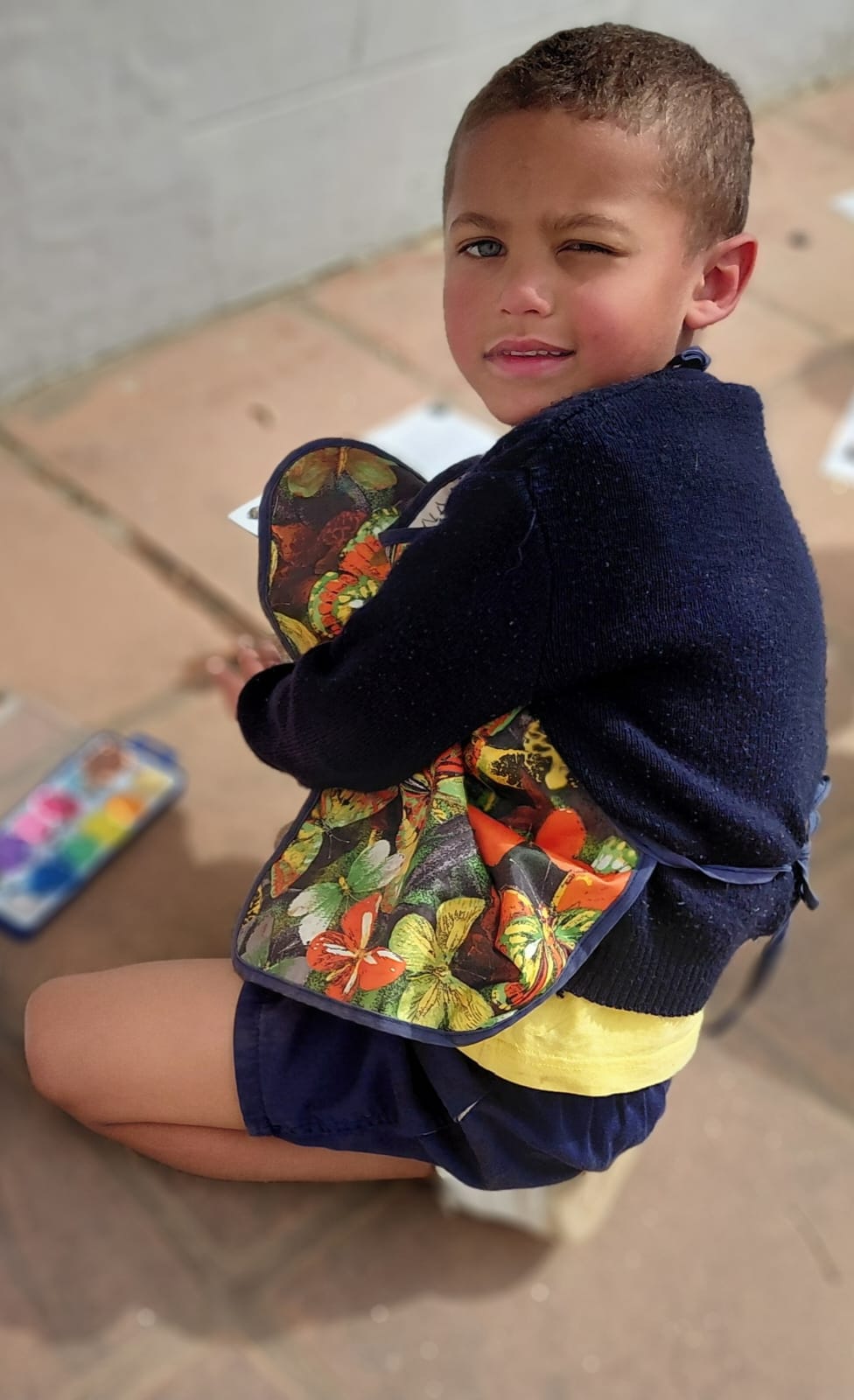 A JENS Grade 1 learner in a butterfly-print apron looking up from his outdoor painting