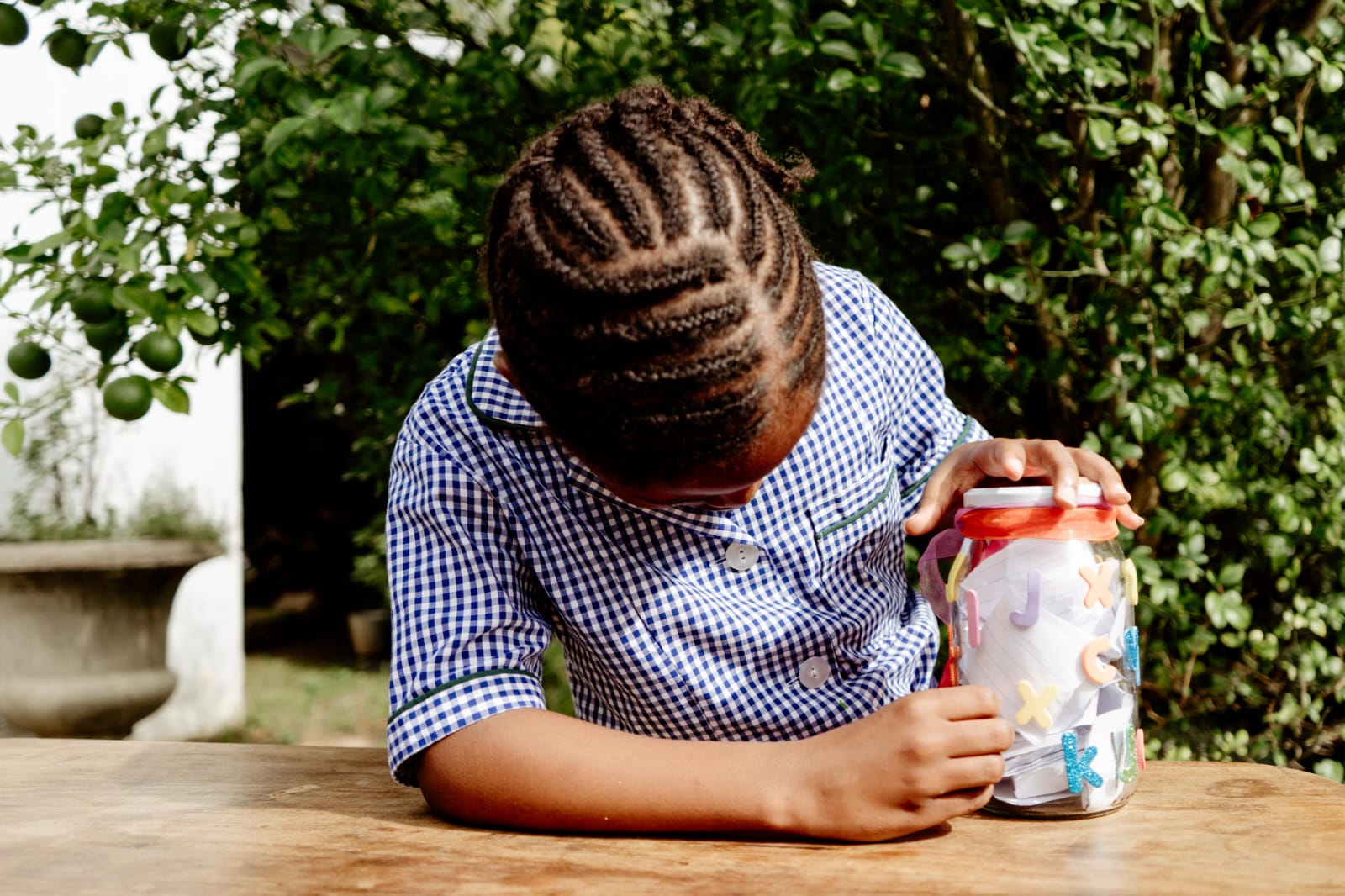 A JENS learner in the checked school dress opening a jar of paper letters at a table in the garden