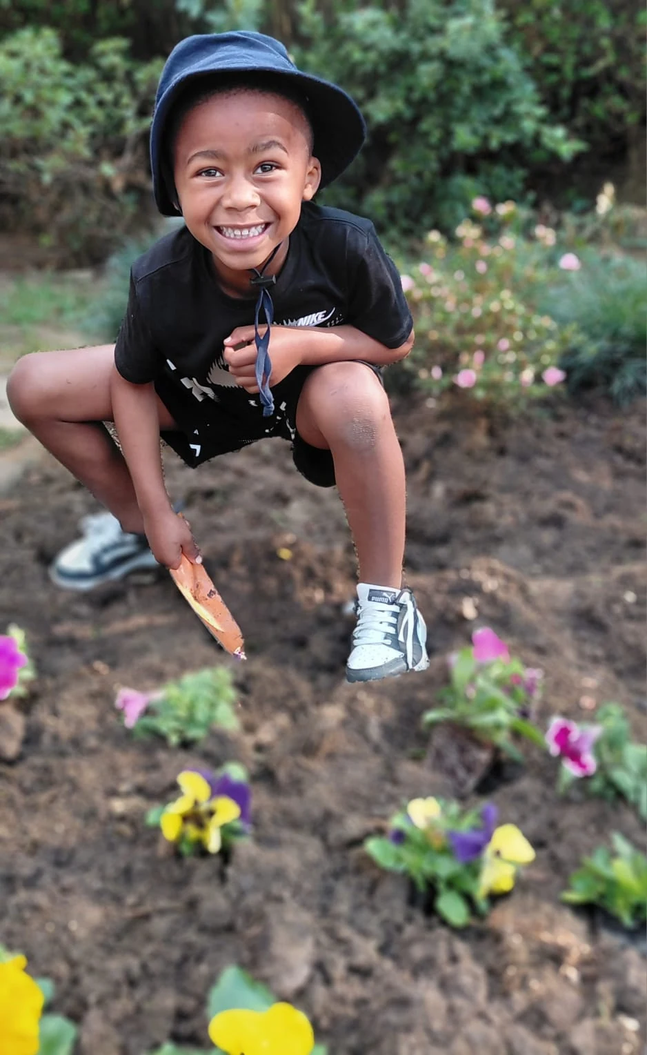 A JENS learner in a sunhat holding a trowel and laughing while planting pansies in a freshly dug bed