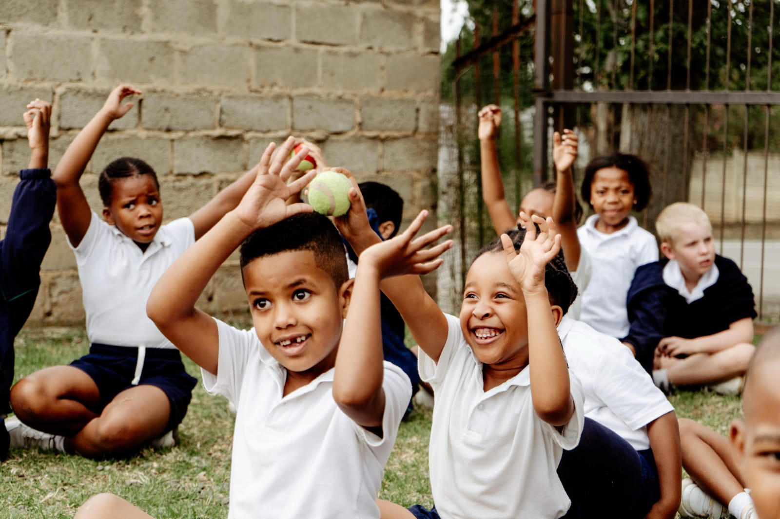 A group of JENS learners sitting on the grass with their hands in the air, holding tennis balls during an outdoor sports lesson