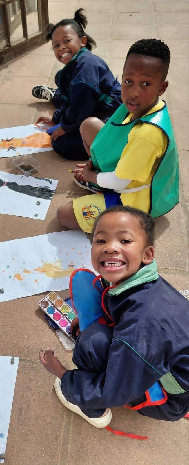 Three smiling JENS Grade 1 learners in painting aprons sitting on the paving with their cat paintings