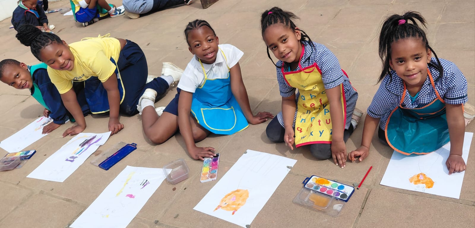 A row of JENS Grade 1 learners in aprons, kneeling on the paving outdoors and painting cats with watercolour paints