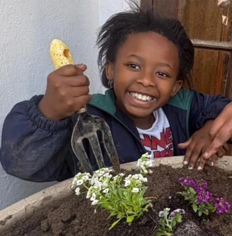 A JENS learner grinning while holding a garden fork above a planter filled with white and purple alyssum