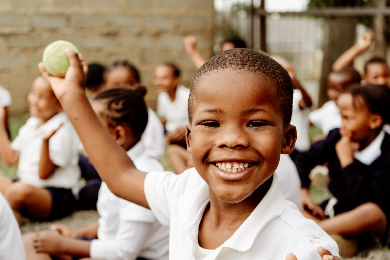 A smiling JENS learner holding up a ball outside, with classmates sitting together behind him